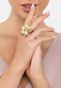 Gold-toned floral ring featuring textured petals and a yellow gemstone center, set on a model's hand with light-colored nails.