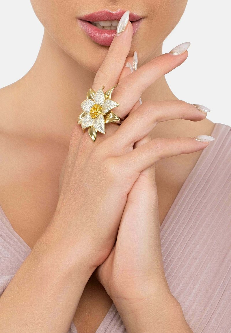 Gold-toned floral ring featuring textured petals and a yellow gemstone center, set on a model's hand with light-colored nails.