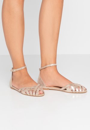 Sandals featuring a beige leather upper with woven design, ankle strap, and flat sole. Smooth texture with metallic accents.