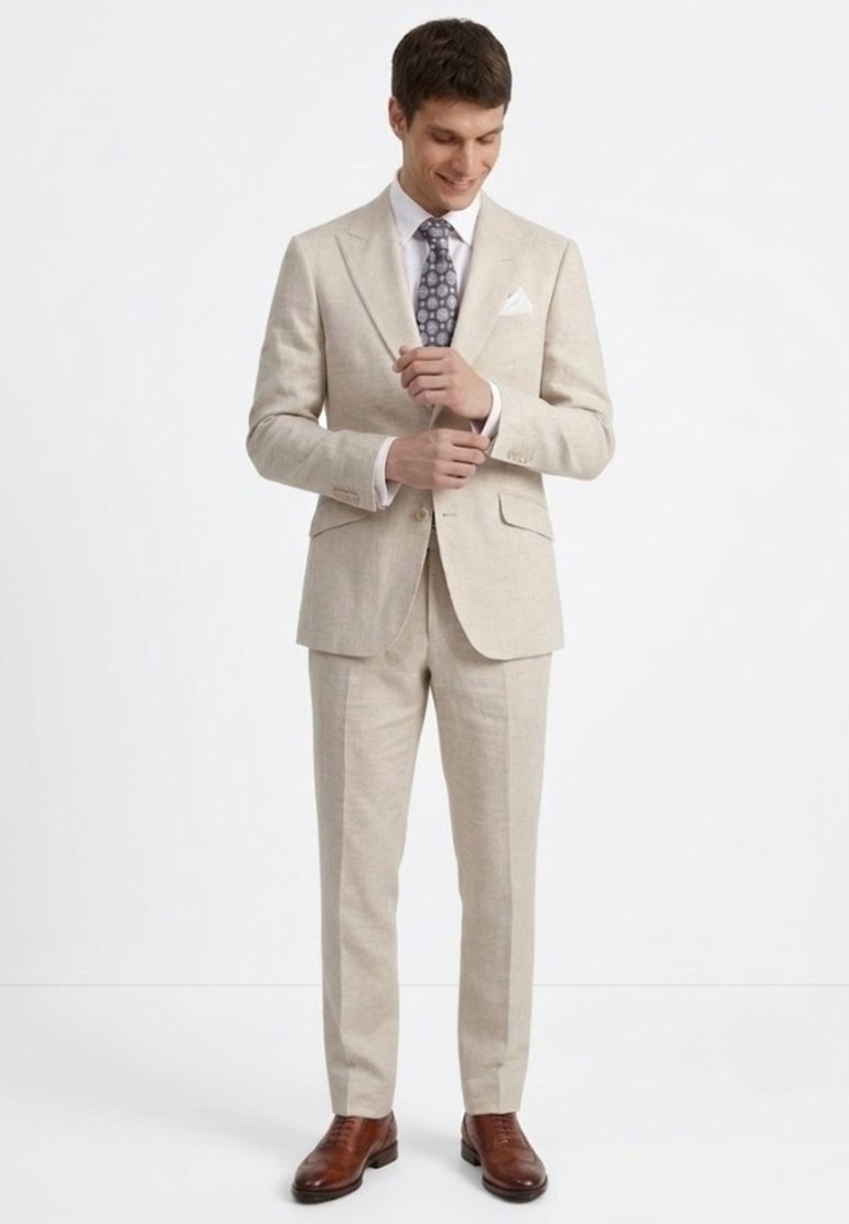 Hombre ajustándose los puños, vestido con traje beige, camisa blanca, corbata con estampado, pañuelo blanco en el bolsillo y zapatos de vestir de cuero marrón, de pie sobre un fondo blanco.