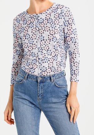 Kvinde iført en langærmet knaplukket blomsterbluse med pink og blå blomster, stukket ned i højtsiddende blå jeans.