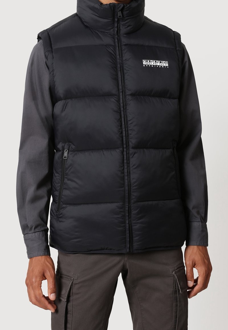 Homme portant une doudoune noire avec un logo Napapijri blanc sur une chemise à manches longues foncée et un pantalon cargo gris, bras détendus.