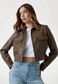 Giacca cropped in pelle marrone con colletto, due tasche frontali e sei bottoni. Indossata sopra una maglietta bianca, abbinata a jeans azzurri chiari.