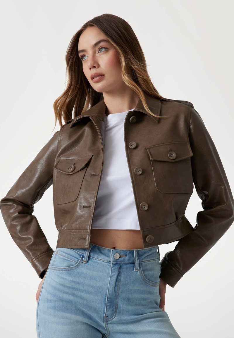 Giacca cropped in pelle marrone con colletto, due tasche frontali e sei bottoni. Indossata sopra una maglietta bianca, abbinata a jeans azzurri chiari.