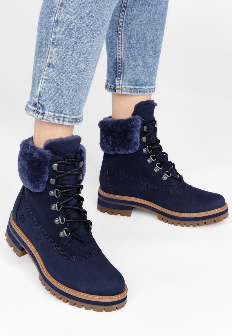 Bottes en daim bleu marine avec un col en fourrure douce, un devant lacé et une semelle en caoutchouc durable, présentant un accent marron et une semelle rainurée.
