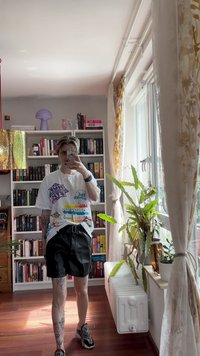 T-shirt grafico bianco con stampe colorate, pantaloni corti neri e sneakers con motivo. Sullo sfondo ci sono librerie e piante d'appartamento.