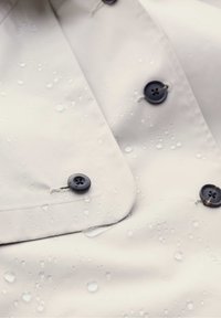 Vaaleanbeige sadetakki, jossa on vedenpisaroita pinnalla. Takissa on tummansiniset napit ja sileä, matta pinta.