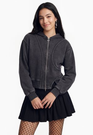 Junge Frau mit langen dunklen Haaren, die einen dunkelgrauen Zip-Hoodie, einen schwarzen Rock, Netzstrumpfhosen und hängende Stern-Ohrringe trägt und leicht lächelt.
