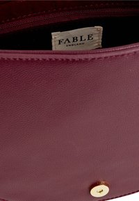 Wnętrze bordowej skórzanej torby o fakturowanym wykończeniu, z materiałową metką "FABLE ENGLAND" oraz złotym zapięciem na guzik.
