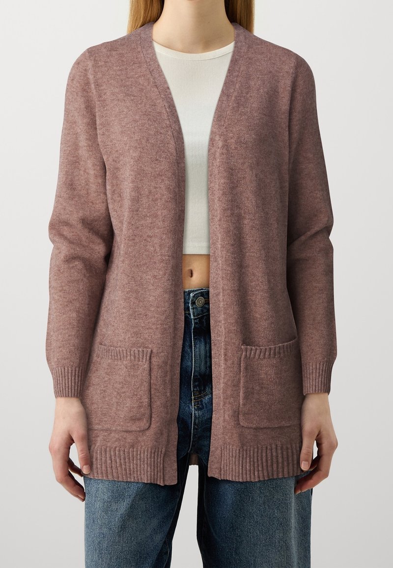 Femme portant un long cardigan mauve avec poches devant, par-dessus un top court blanc côtelé et un jean bleu sur un fond uni.