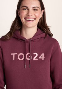 Young woman smiling, wearing a maroon hoodie with "TOG24" logo in light pink letters on a plain light background.