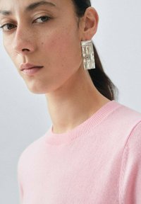 Rosa tröja med rund halsringning, med en slät textur. Stora geometriska genomskinliga örhängen med silverdetaljer.
