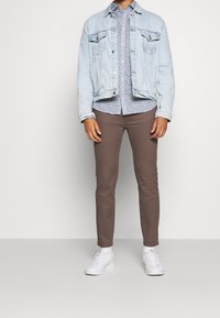 Ljusblå jeansjacka över en blommig skjorta, bruna slim-fit byxor och vita sneakers. Släta texturer och avslappnad passform.