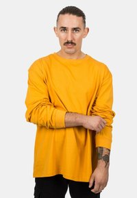 Langärmliges, Rundhals-T-Shirt aus leuchtend gelbem Baumwollstoff. Merkmale sind der lockere Schnitt und hochgerollte Ärmel, getragen über schwarzen Hosen.