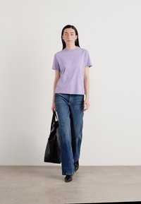 Lila crew-neck t-paita hienovaraisella brodeerauksella, yhdistettynä leveälahkeisiin sinisiin farkkuihin ja mustaan nahkalaukkuun, neutraalilla taustalla.
