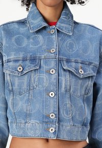 Veste en denim de couleur bleu clair, au design court, avec deux poches poitrine et des fermetures à boutons argentés ornées de motifs de logo subtils.