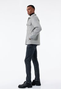 Chaqueta de punto gris con dos bolsillos frontales, llevada sobre un suéter de cuello alto negro, combinada con jeans oscuros y zapatos de cordones negros, de pie de lado.
