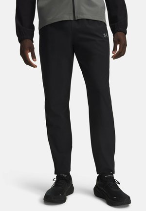 Onderlichaam van een persoon die zwarte Under Armour-broeken, zwarte sneakers en een grijs jack met zwarte mouwen draagt tegen een witte achtergrond.