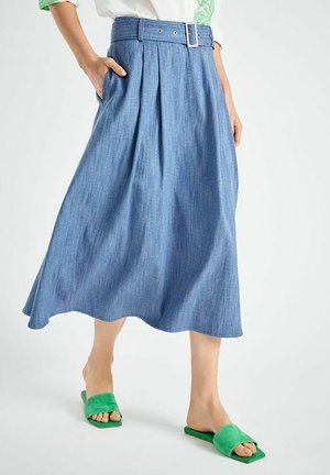 Denim-Midirock mit Falten, Gürtelschlaufen und breitem Gürtel; hellblaue Farbe mit subtiler Textur. Kombiniert mit grünen flachen Pantoletten.