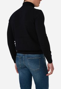 Schwarzer Rollkragenpullover mit geripptem Kragen und Bündchen, taillierte Passform. Getragen mit blauen Denim-Jeans, die über gebogene Gesäßtaschen verfügen.