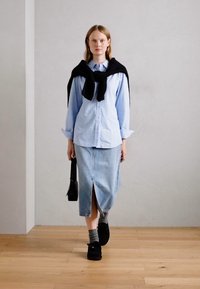 Femme marchant à l'intérieur portant une chemise bleu clair, une jupe midi en denim avec une fente devant, des chaussures noires, des chaussettes grises et un pull noir sur les épaules.