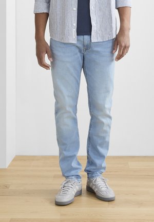 Lichtblauwe jeans met een vervaagde afwerking, slim fit en een gemiddelde taille, gecombineerd met lichtgrijze sneakers. Houten vloer als achtergrond.
