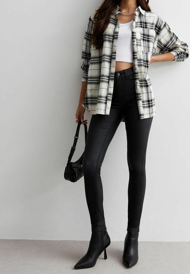 Femme portant un legging noir taille haute, des bottines noires à talon pointu, un crop top blanc, une chemise oversize à carreaux noir et blanc, tenant un petit sac noir.