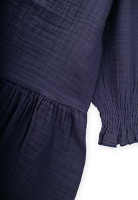 Tumesinine kangas, millel on kogutud elastsed mansetid ja volangid pikadel varrukatel valgel taustal.