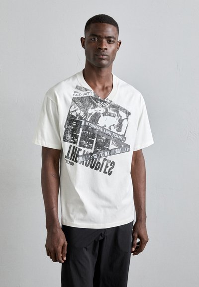 T-shirt bianca a maniche corte con scollatura a V, caratterizzata da stampe grafiche nere di testi e immagini, tra cui "ESaurito!" e "TOUR DELLA VITA ETERNA."