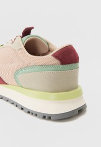 Multicolored sneaker featuring a mesh upper in beige, red suede accents, a green rubber overlay, and a chunky white and gray sole.