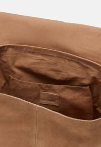 Wnętrze brązowej torby z zamszu, z miękkim materiałowym wykończeniem, małą wewnętrzną kieszonką i skórzaną metką. Gładka tekstura na całej powierzchni.