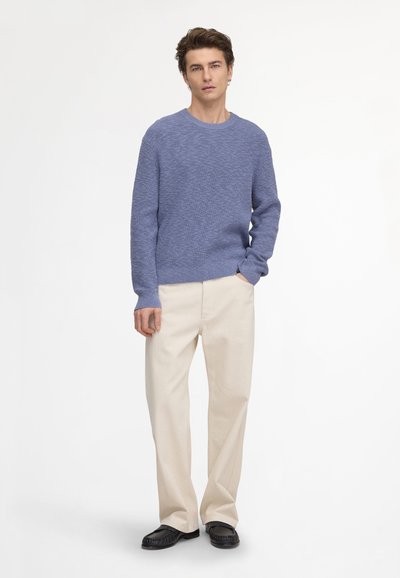 Jeune homme portant un pull en maille bleu, un pantalon couleur crème et des mocassins noirs, debout avec une main dans la poche sur fond blanc.
