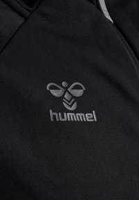 Zwarte sportieve stof met een geborduurd grijs logo met een abstract ontwerp en daaronder het woord "hummel". Gladde textuur.