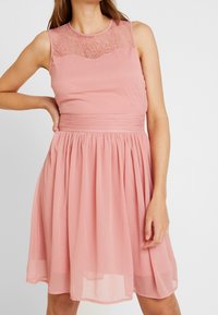 Robe rose avec un corsage en dentelle, design sans manches et jupe plissée. Tissu lisse avec une taille cintrée et une silhouette fluide.