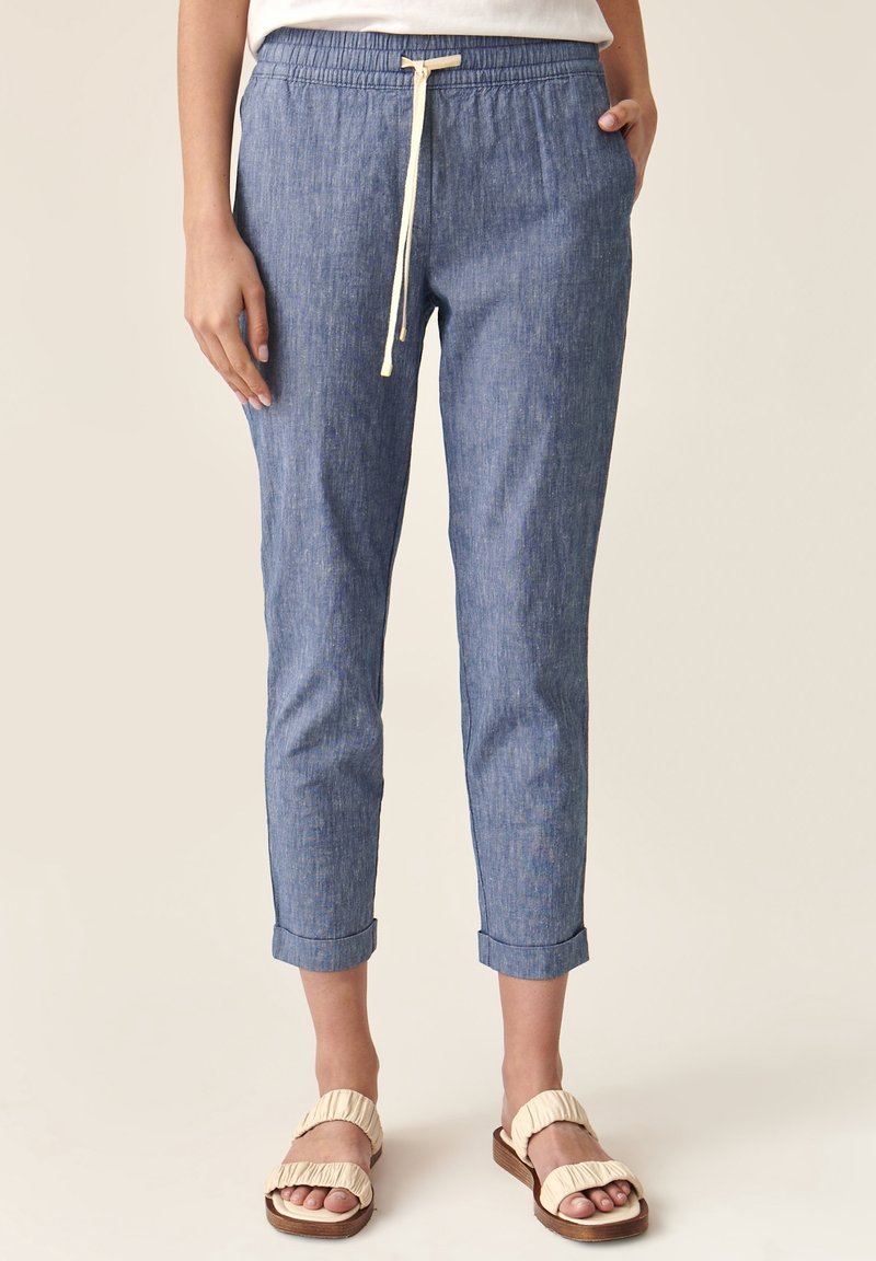 Femme portant un pantalon en jean bleu retroussé avec une taille élastique et un cordon blanc, associé à des sandales beiges sur un fond uni.
