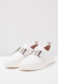 Vita sneakers med elastiska remmar, metalliska logotypdetaljer, slät konstläderövre och gummisålar. Minimalistisk design utan synliga snören.