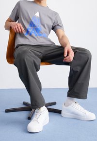 Graues Baumwoll-T-Shirt mit blauem Berggrafik, kombiniert mit dunklen Jogginghosen und weißen Turnschuhen. Model sitzt auf einem Holzstuhl vor einem schlichten Hintergrund.