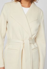 Abrigo cruzado color crema hecho de tela suave, con un cuello amplio, cinturón para anudar y dos bolsillos delanteros. Textura lisa con costuras cuidadas.