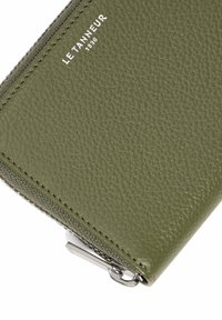Portefeuille en cuir vert olive avec une surface texturée, fermeture éclair argentée et branding en relief. Forme compacte et rectangulaire.