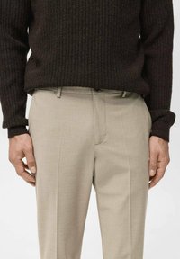 Pantalones de traje beige con una textura suave, pliegue frontal y trabillas para cinturón. Combinando con un suéter de canalé oscuro, manos descansando en la cinturilla.