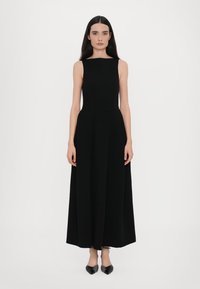 Zwarte mouwloze maxi-jurk met een aansluitend bovenlijfje, uitlopende rok en een soepele textuur. Gestikte taille detail, gedragen met puntige zwarte schoenen.