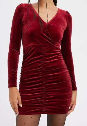 Frau trägt ein figurbetontes rotes Samtkleid mit langen Ärmeln und gerafften Details, kombiniert mit schwarzen, transparenten Strumpfhosen mit kleinen Polka-Dots.