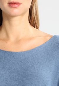 Hellblaues, geripptes Strickoberteil mit weitem Bootsausschnitt. Weiche Textur mit sichtbaren Nähten und subtiler Glanz.