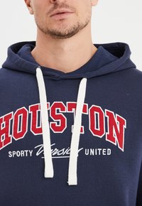 Felpa blu navy con cappuccio, caratterizzata da cordini bianchi, grande testo rosso "HOUSTON" e "SPORTY VARSITY UNITED" in bianco. Tessuto morbido.