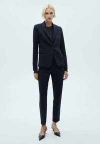 Costume tailored navy avec une veste à un bouton, un pantalon droit et des talons noirs pointus. Tissu lisse avec un design élégant et professionnel.