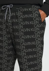 Czarne spodnie dresowe z powtarzającym się szarym wzorem logo "CALVIN KLEIN", z elastycznym pasem ze splecionym sznurkiem i kieszeniami po bokach.