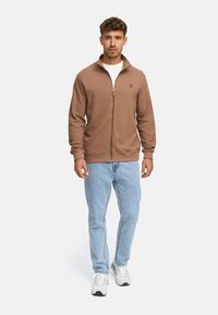 Hombre con chaqueta marrón de cremallera, camiseta blanca, jeans azul claro y zapatillas blancas, de pie contra un fondo liso.