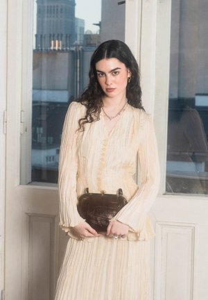 Kvinde med langt mørkt hår iført en cremefarvet plisseret kjole, der holder en mørkebrun tekstureret håndtaske, stående indenfor ved glasdøre.