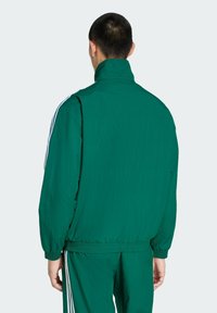 Zielona kurtka przeciwwiatrowa z wysokim kołnierzem, z białymi paskami po bokach, elastycznymi mankietami i luźnym krojem. Materiał wydaje się lekki i gładki.