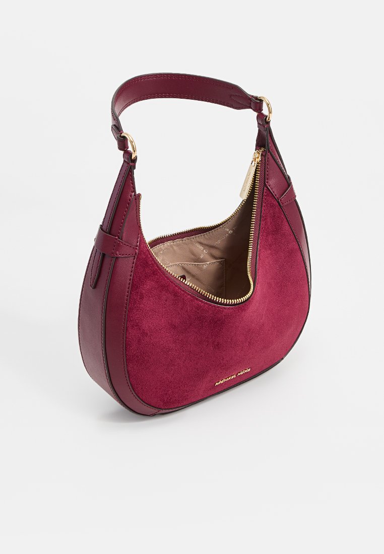 Sac à main en daim bordeaux ayant une forme courbée, fermeture éclair dorée et une petite poignée. L'intérieur présente une doublure beige. La marque est visible sur le devant.